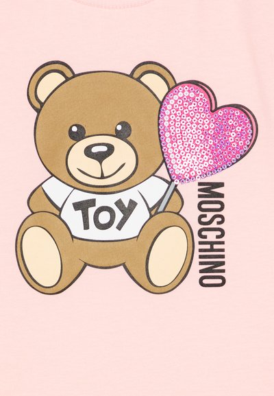 Gráfico de un oso de peluche marrón con un globo corazón de lentejuelas rosas, vistiendo una camiseta blanca con la palabra "JUGUETE" impresa sobre un fondo rosa claro.