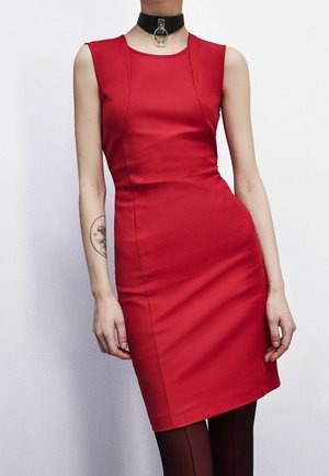 Mouwloze rode nauwsluitende jurk op een staand persoon met een zwarte chokerketting en een ronde tatoeage op de linker onderarm.