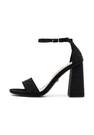 Sandalía negra con textura de ante, una correa en el tobillo, un ancho tacón bloque adornado con pequeñas tachuelas y una correa rectangular para los dedos.