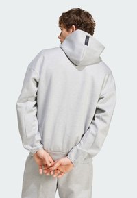 Svetlo šedá mikina s kapucňou, vyrobená z textúrovanej látky, vo voľnom strihu, so malou štítkom na zadnej strane. Model má ruky zložené.