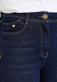 Los vaqueros de mezclilla azul oscuro presentan costuras doradas, un bolsillo delantero con un remache de metal y un botón decorativo en la cintura.