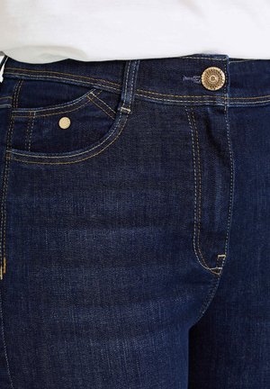 Un jean en denim bleu foncé avec des coutures dorées, une poche avant ornée d'un rivet métallique, et un bouton décoratif à la taille.