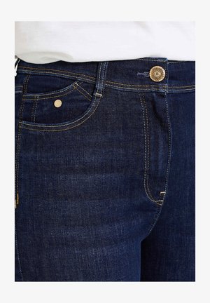 Los vaqueros de mezclilla azul oscuro presentan costuras doradas, un bolsillo delantero con un remache de metal y un botón decorativo en la cintura.