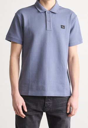 Personne portant un polo à manches courtes de couleur violet clair avec le logo Calvin Klein sur la poitrine et un jean gris foncé, se tenant contre un fond uni.