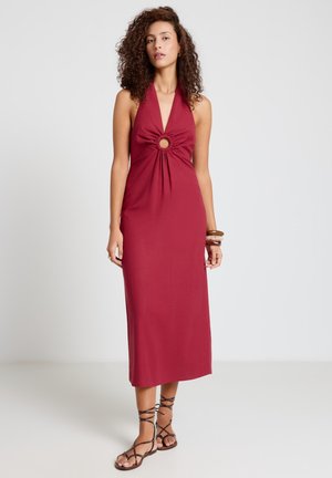 Femme aux cheveux bouclés portant une robe midi rouge sans manches à col noué avec un détail d'anneau à l'avant et des sandales marron à lanières sur un fond uni.