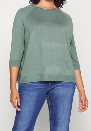 Persoon draagt een loszittend saliegroen shirt met driekwart mouwen en een blauwe spijkerbroek, staand tegen een effen lichte achtergrond.