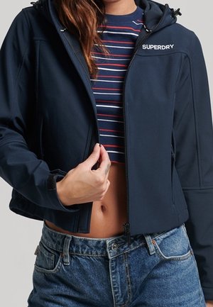 Femme portant une veste Superdry bleu marine par-dessus un crop top rayé et un jean bleu taille haute, ajustant la fermeture éclair de la veste.