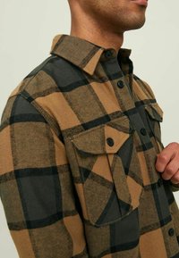 Uomo che indossa una camicia di flanella a scacchi marrone e nera con tasche sul petto bottate, mentre sistema i bottoni anteriori.
