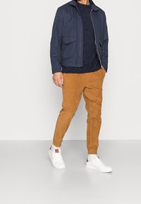 Navy-Jacke mit Reißverschluss und zwei Fronttaschen, getragen über einem strukturierten navyblauen Pullover, kombiniert mit beigen Cordhosen und weißen Turnschuhen.
