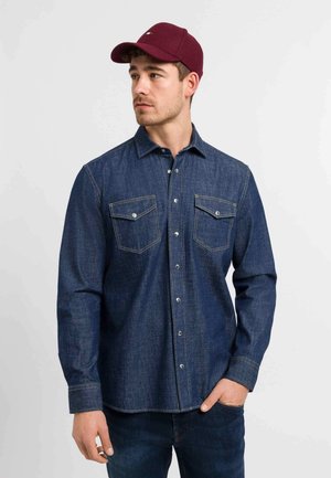 Donkerblauwe denim shirt met lange mouwen, twee borstzakken, knopen aan de voorkant en contrasterende gele stiksels. Gecombineerd met een bordeauxrode pet.