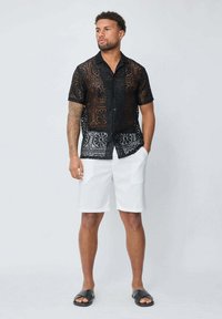 Chemisier noir en dentelle à manches courtes avec une texture à motifs, associé à un short blanc. Le mod èle se tient sur un fond uni, portant des sandales.