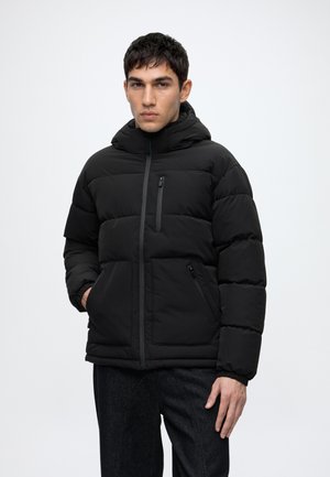 Jeune homme portant une veste matelassée noire à capuche avec poches zippées et un pantalon noir, debout devant un fond clair uni.