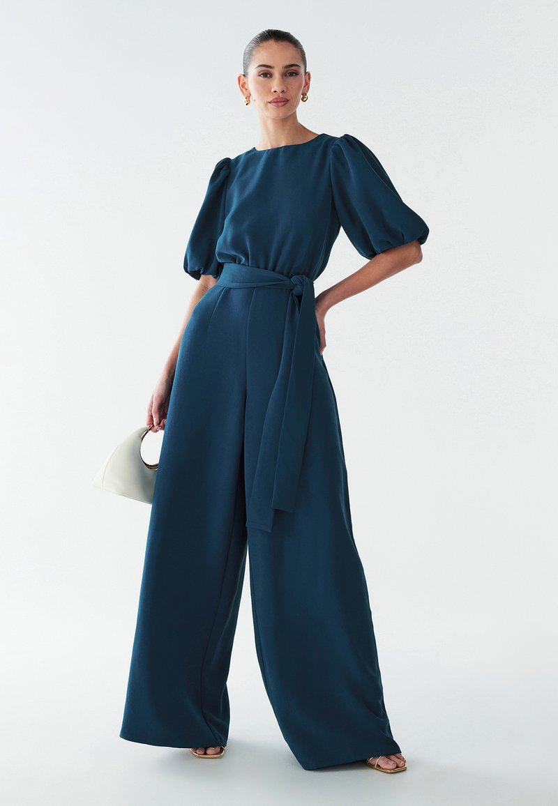 Turquoise jumpsuit met pofmouwen en een ceintuur in de taille. Wijd uitlopende pijpen, gladde stof, geaccentueerd door een beige handtas en metallic sandalen.