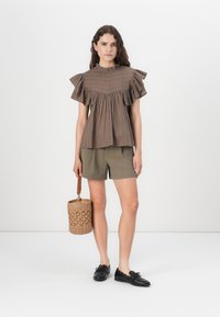 Bruine, gerimpelde top met verticale strepen, gecombineerd met olijfgroene shorts. Het model houdt een ronde, geweven tas vast en draagt zwarte schoenen.
