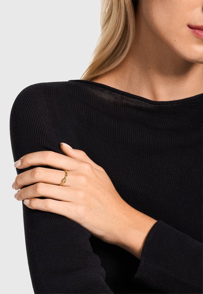 Frau in schwarzem Pullover mit verschränkten Armen, die einen zarten Goldring mit kleinem Knoten-Design an ihrer linken Hand präsentiert.