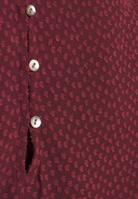 Oberteil aus burgunderrotem Stoff mit strukturierten Punkten, ausgestattet mit einer Knopfleiste und einem Seitenschlitz. Das Material wirkt leicht und hat eine weiche Oberfläche.