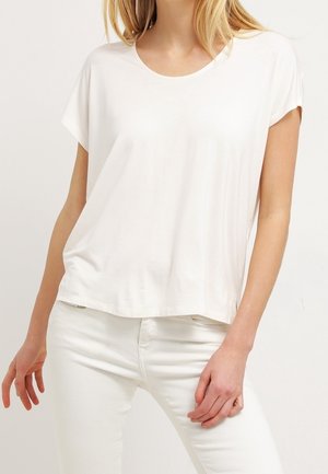 Nainen, joka pukeutunut yksinkertaiseen valkoiseen lyhythihaiseen t-paitaan ja valkoisiin housuihin, seisoo yhdellä kädellä reidellään.