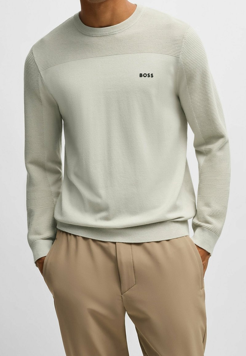 Homme portant un pull côtelé beige clair avec le logo "BOSS" sur la poitrine et un pantalon beige, les mains dans les poches, sur un fond uni.