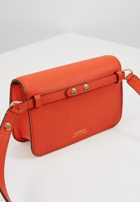 Pomarańczowa skórzana torba na ramię z złotymi elementami, z fakturowaną powierzchnią, zamknięciem na klapę i regulowanym paskiem. Logo wytłoczone na przodzie.