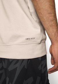 Light beige DRI-FIT athletic shirt with a smooth texture, featuring a stitched hem and a logo near the bottom seam. Worn with patterned dark shorts.