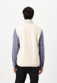 Kremfarget fleece vest med grønn krage, båret over en blå rutete skjorte. Vesten har en avslappet design og en glatt tekstur.