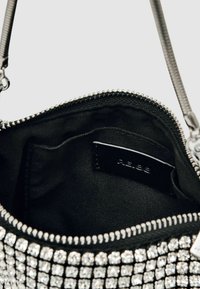 Črna torbica z bleščečim srebrnim zadrgo, prekrita z majhnimi bleščečimi kristali. Notranjost je podložena z črnim tekstilom in ima majhno oznako.