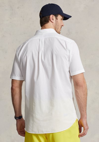 White short-sleeve shirt with a textured finish, button-down collar, and a curved hem, worn with yellow shorts and a navy cap.