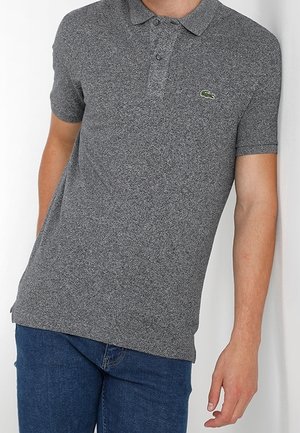 Homme portant un polo gris à manches courtes avec un petit logo crocodile au niveau de la poitrine, associé à un jean bleu, debout contre un mur blanc.