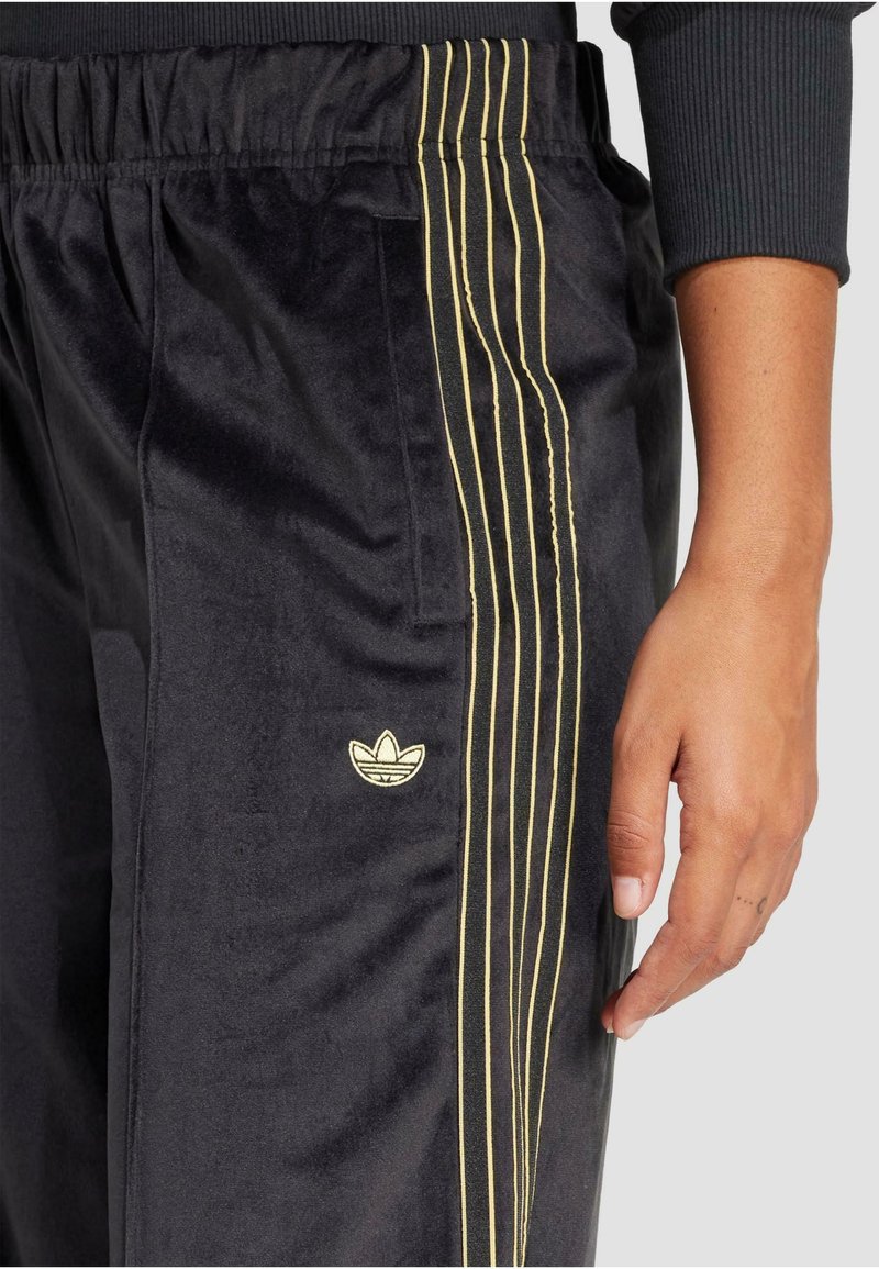 Pantaloni sportivi in velluto nero con una finitura testurizzata, caratterizzati da tre strisce dorate lungo il lato e un logo dorato vicino alla tasca.
