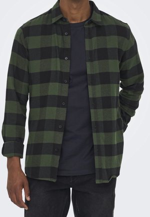 Chemise en flanelle à carreaux verts et noirs avec un devant à boutons, des manches longues et une texture douce, portée par-dessus un T-shirt bleu marine.