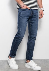 Jean en denim bleu avec une coupe droite, légère usure, design à cinq poches et poignets retroussés, associés à des baskets blanches.