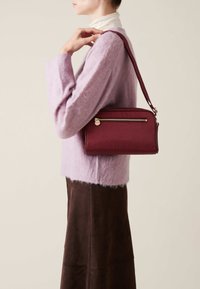 Burgundy leather handbag with a textured finish, featuring a gold-tone zip closure and a long strap, carried by a person wearing a lilac sweater.