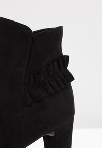 Černé semišové kotníkové boty s volánkovým detailem po boku, se strukturovaným svrškem a vysokým podpatkem. Hladká textura dominuje.