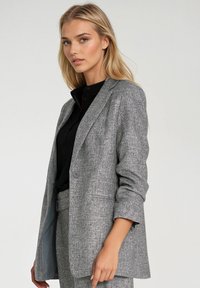 Szary żakiet o strukturalnej fakturze z dopasowanym krojem, z pojedynczym zapięciem na guzik i lekko podwiniętymi rękawami, w połączeniu z czarną koszulą z kołnierzykiem.