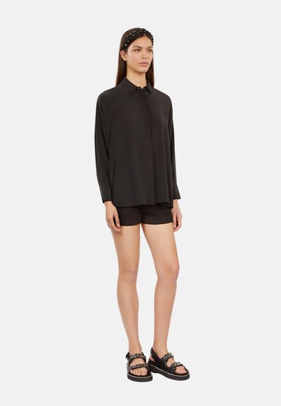 Zwarte blouse met lange mouwen en kraag, gecombineerd met donkergekleurde shorts en zwarte sandalen met enkelbandjes en studs. Glad, lichtgewicht materiaal.