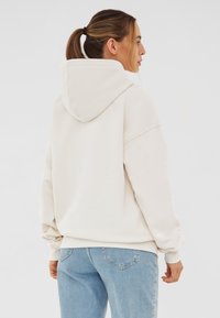 Sudadera color crema hecha de tela suave, con una capucha grande y puños de canalé. Usada con jeans azul claro y un corte relajado.