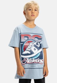 Blond dziecko nosi jasnoniebieską, oversize'ową koszulę z dużą grafiką trampka oraz napisem "DC Athletics" na przodzie.