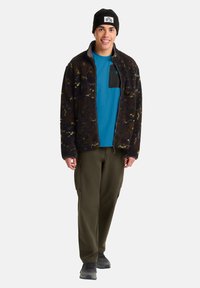 Fleecejacke in Dunkelbraun mit gelben Akzenten, schwarzer Tasche; blaues Hemd darunter; olivgrüne Cargo-Hose; schwarze Mütze; graue Sneakers.