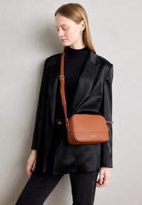 Brown leather crossbody bag with a rectangular shape, textured finish, and adjustable strap, worn with a black jacket and top.