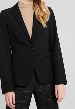 Femme portant un blazer noir à un bouton sur un col roulé côtelé beige et un pantalon à carreaux noir avec des lignes brunes et bleues.