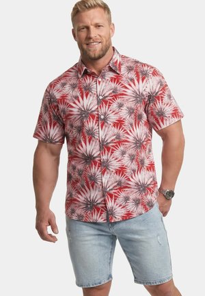 Mann lächelt, trägt ein rotes kurzärmeliges Hemd mit Blumenmuster und hellblaue Jeans-Shorts mit einer Armbanduhr am linken Arm.