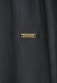 Tela negra con una superficie texturizada que presenta una etiqueta metálica en tono dorado con la inscripción "D'NEILL" adherida. Diseño simple y minimalista.