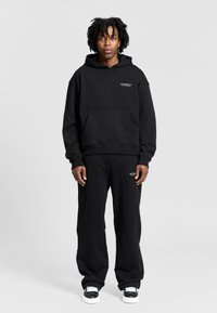 Black hoodie with kangaroo pocket and logo on the chest, paired with matching black sweatpants and black sneakers with white accents.