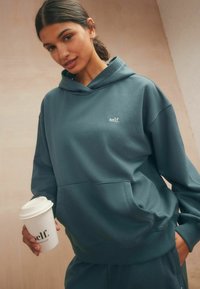 Übergroßer türkisfarbener Hoodie mit einer Vorderseite-Tasche und weißem "self." Logo. Das Modell hält einen weißen Kaffeebecher. Glatte Textur, lässiges Design.