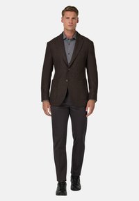 Brązowa, strukturalna marynarka z klapami, dwoma przednimi kieszeniami, zestawiona z szarym polo i ciemnymi spodniami. Czarnym butom nadaje całości elegancji.