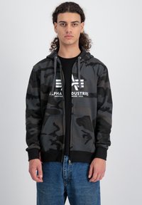 Kamuflažna zip-up dukserica u nijansama tamno sive i crne boje s kapuljačom na šnuranje, prednjim džepovima i istaknutim bijelim logom. Nosi se preko crne majice.