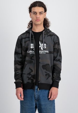 Sudadera con cremallera de camuflaje en tonos de gris oscuro y negro, con capucha ajustable, bolsillos frontales y logotipo blanco prominente. Lleva puesta una camiseta negra debajo.