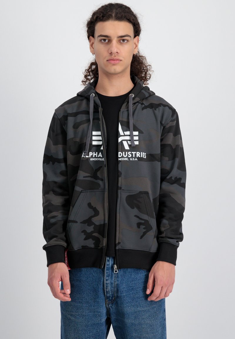 Kamuflažna zip-up dukserica u nijansama tamno sive i crne boje s kapuljačom na šnuranje, prednjim džepovima i istaknutim bijelim logom. Nosi se preko crne majice.