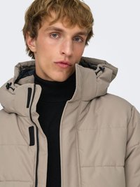 Puffer jacket with a beige exterior, black accents, hood, and zip front. Features quilted texture and high collar.