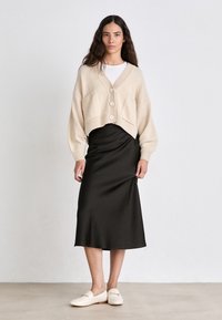 Beige cropped cardigan with ribbed cuffs, four buttons, and pockets, paired with a black satin midi skirt and cream loafers on a neutral surface.